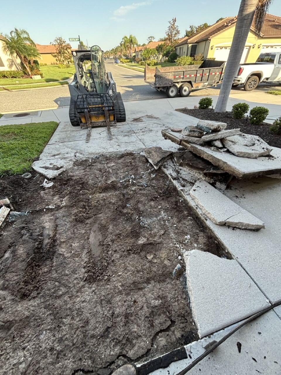 Compact loader beside broken concrete driveway slabs with a hauling trailer parked at the curb